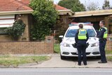Four police officers are outside a brick house. Two officers stand in front of a police car, while one man is in a forensic suit