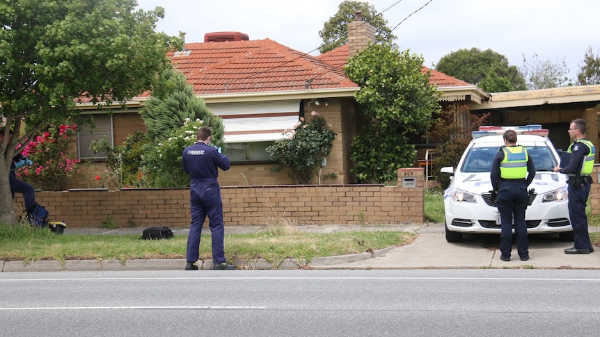 Four police officers are outside a brick house. Two officers stand in front of a police car, while one man is in a forensic suit