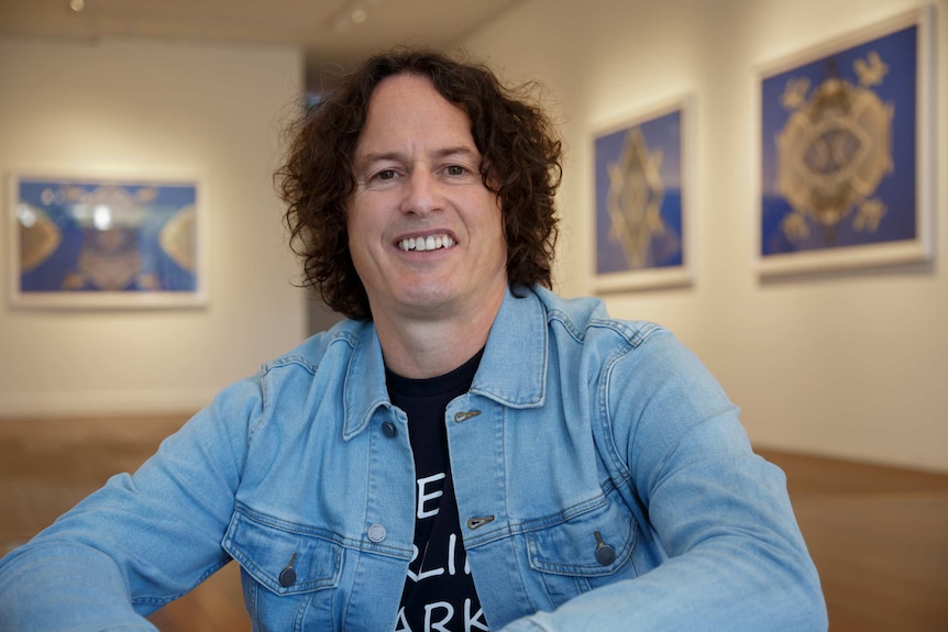 The artist Kent Morris smiling for the camera, his vivid blue artworks hanging in the background in a gallery