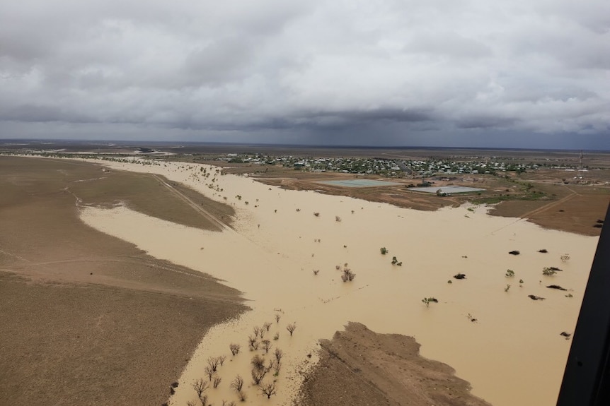 An aerial shot of floodwaters with trees poking out. On one side of the floodwaters is a town; on the other, a flat expanse.