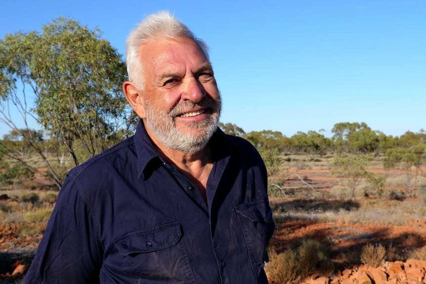A man smiles to the camera, behind him is red dirt and some bushy scrub.
