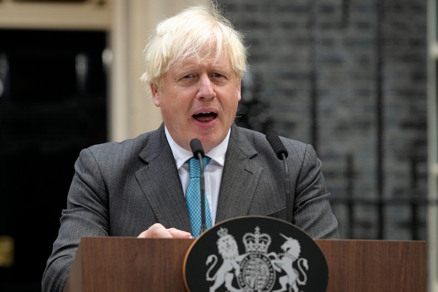 An older man wearing a blue tie stands at a lectern outside number 10.