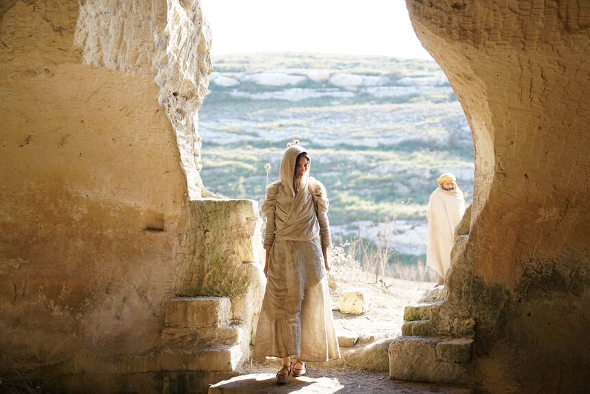Colour still image from 2018 film Mary Magdalene entering a cave on a sunny day, wrapped head to toe in cloth.