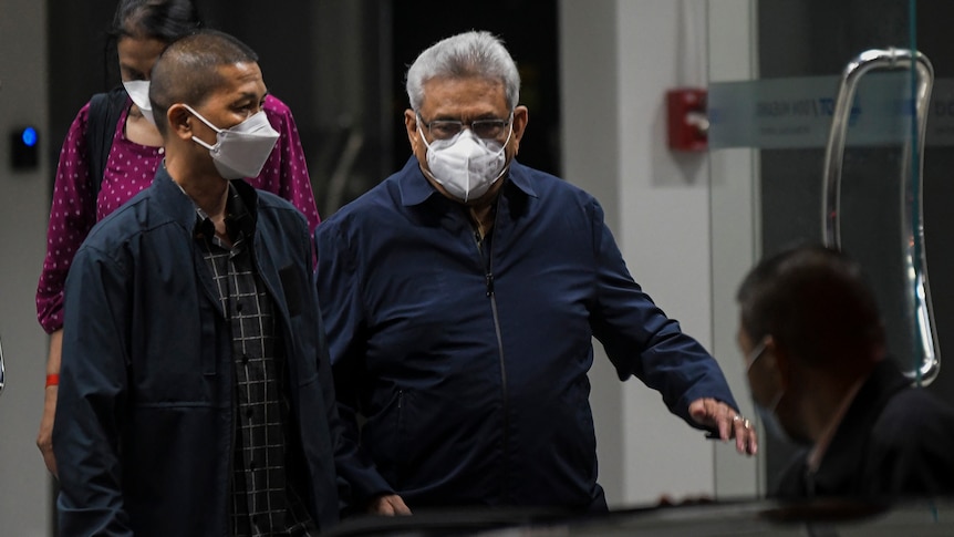 A white-haired Sri Lankan man with a blue jacket and wearing an N95 respirator mask walks towards a waiting car.