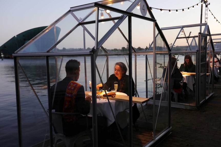 A couple sitting in a glass enclosure with the water in the background and candles as they eat.