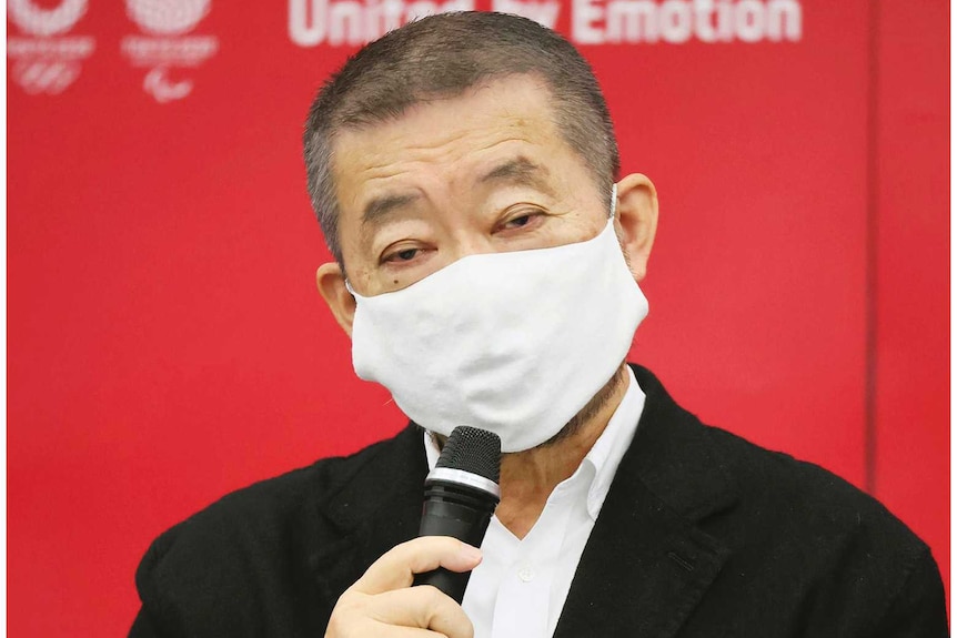 Hiroshi Sasaki speaks into a microphone in front of a red background, wearing a mask