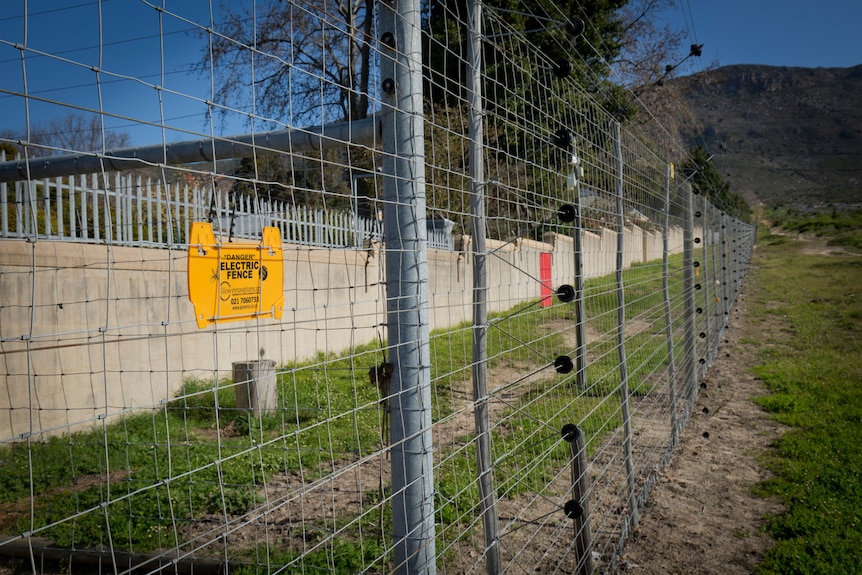 A high, electrified fence runs along the edge of a residental area towards a mountain.