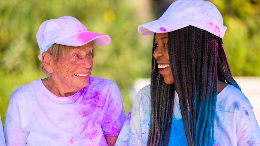 An elderly white woman and a Black teenager both wearing white t-shirts and caps that have been stained with bright colours.