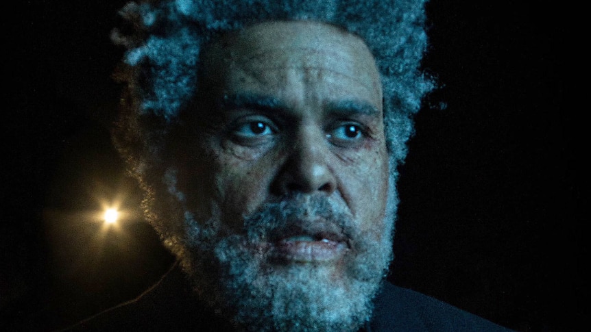The Weeknd with prosthetic make up to make him look elderly, with light shining in the background