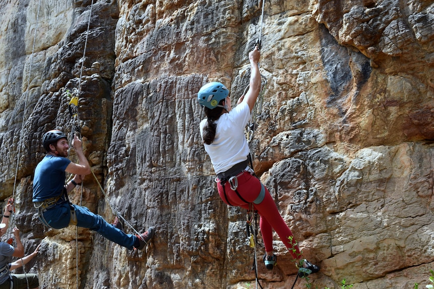 A female rock climber moves up a natural rock face while someone next to her watches.