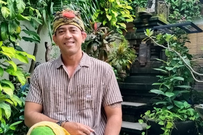 A man in a traditional Balinese headdress sits in a tropical garden