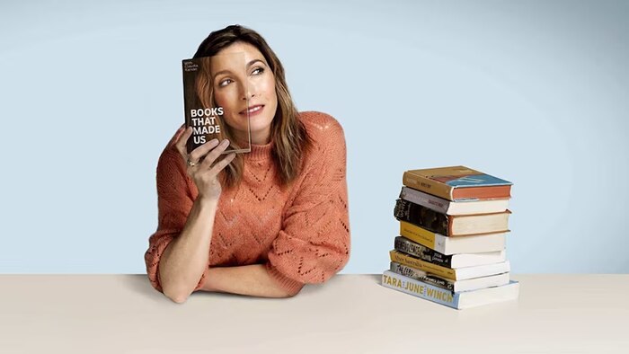 Woman holding a book with half her face on it, covering half her face and a pile of books next to her on the table she sits at.