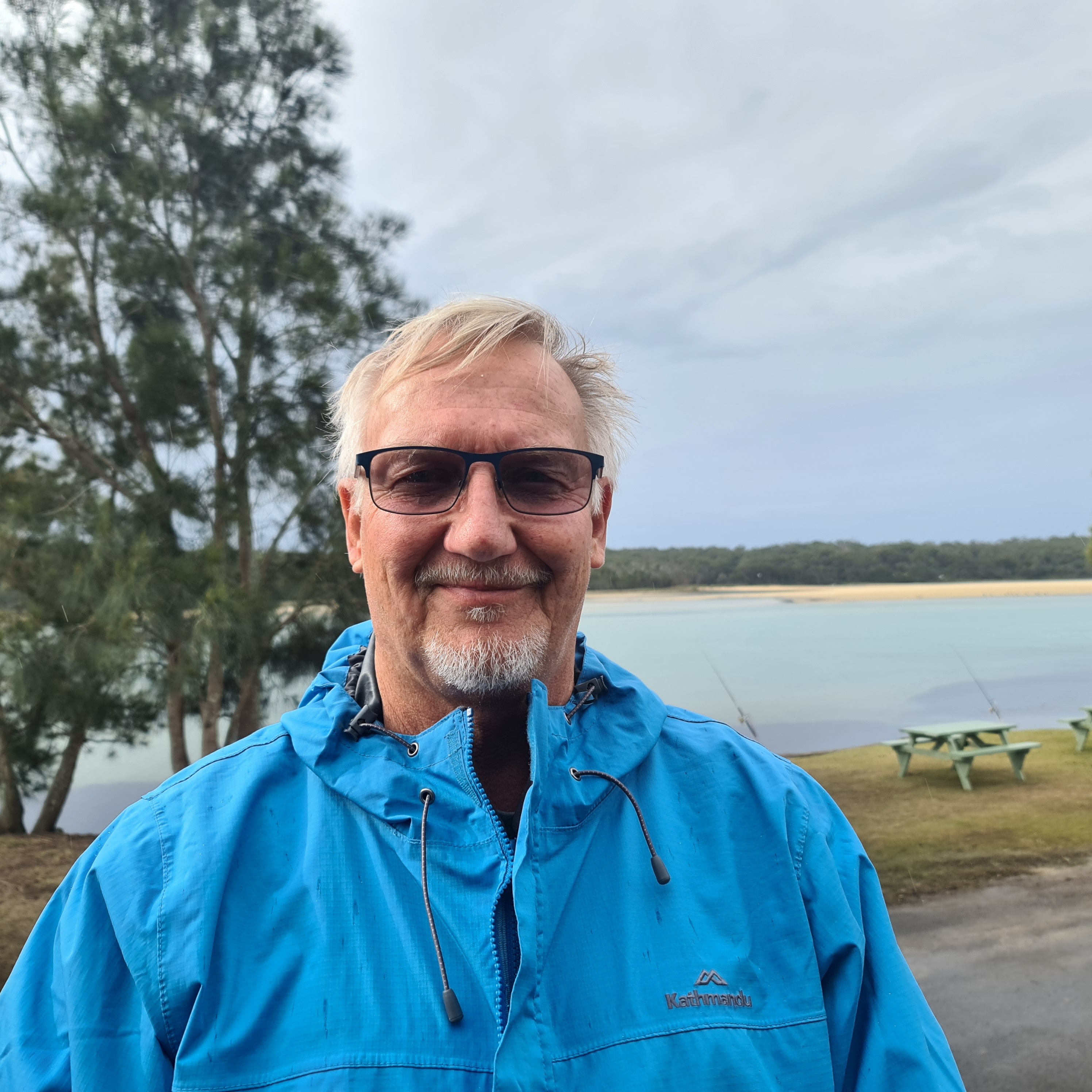 A smiling, bespectacled man with gray hair and a neat beard in front of a waterway, wearing a rain jacket.