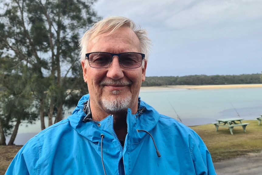 A smiling, bespectacled man with gray hair and a neat beard in front of a waterway, wearing a rain jacket.