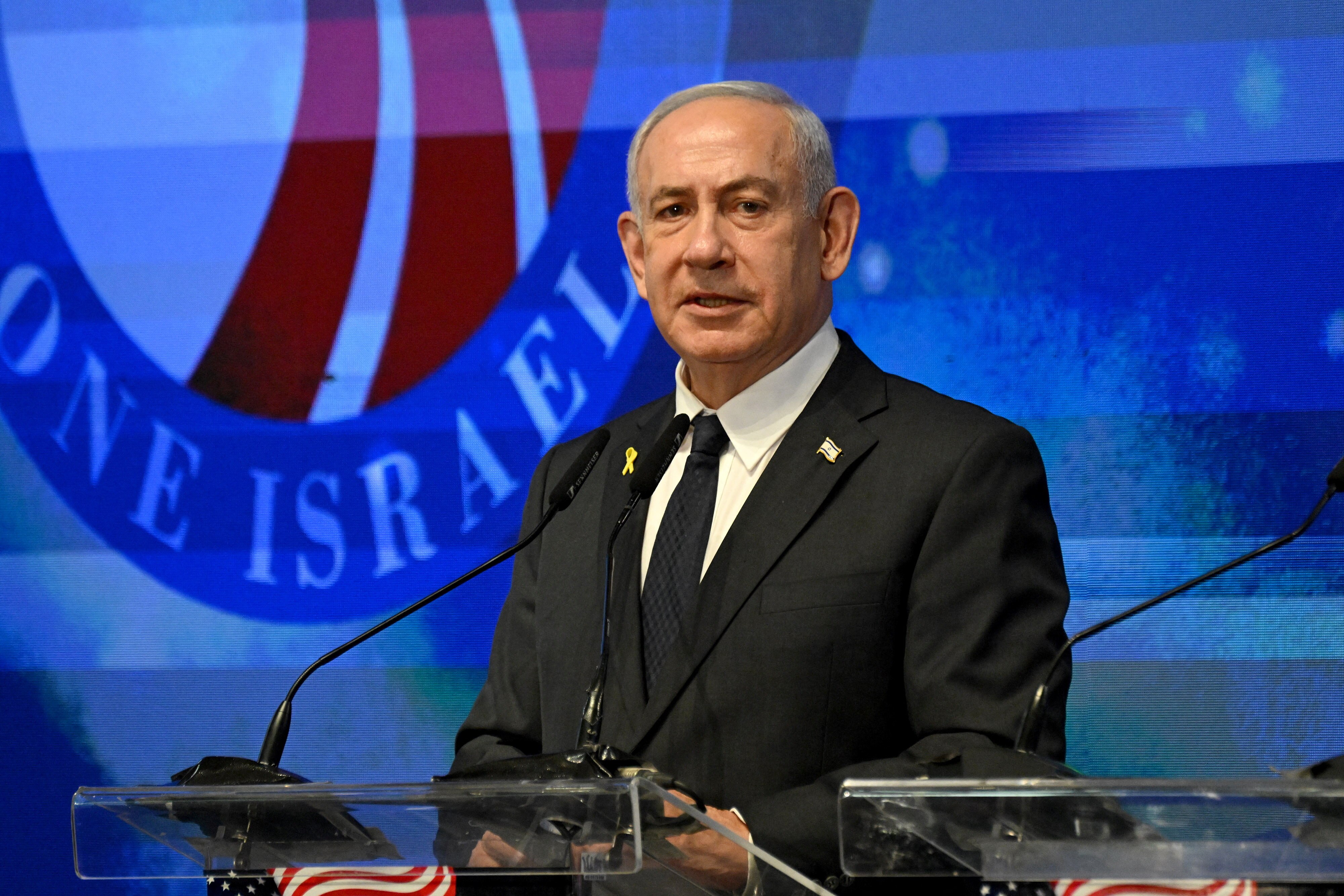 A man wearing a black suit an an Israeli flag pin speaks on a stage
