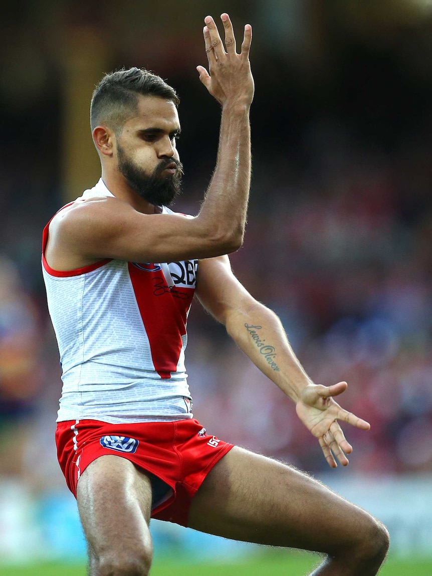 Sydney Swans player Lewis Jetta performing an Aboriginal dance with his arms on a football field.