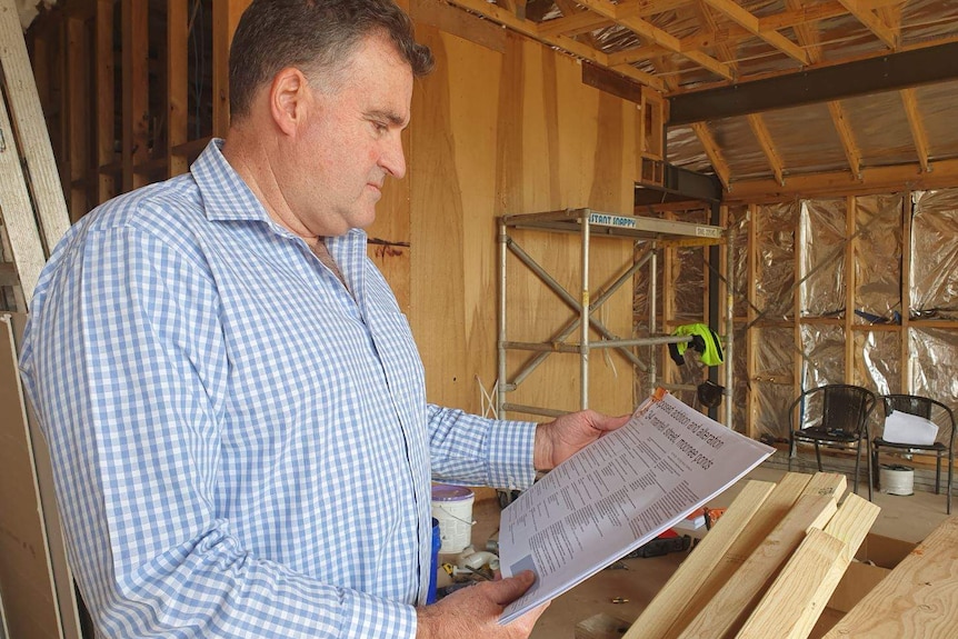 A picture of a man in a business shirt, looking at plans, inside a house being built, with the frames visible.