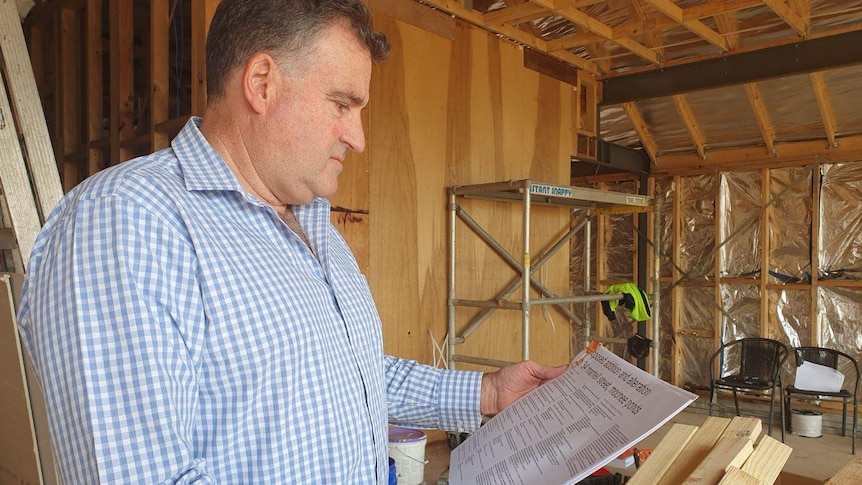 A picture of a man in a business shirt, looking at plans, inside a house being built, with the frames visible.