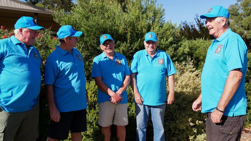Five men standing in front of a bush, all five men are wearing a blue shirt and matching blue hat.