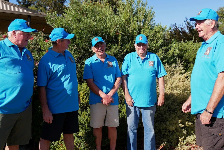 Five men standing in front of a bush, all five men are wearing a blue shirt and matching blue hat.