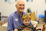 An elderly man holds a red headed toddler on his lap and smiles into the camera