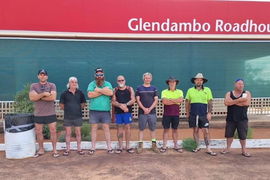 8 men stand outside the Glendambo roadhouse sign with crossed arms looking frustrated.