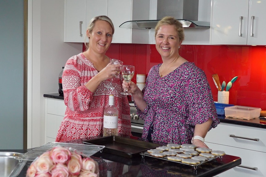 Two women smile and clink their wine glasses in a kitchen.