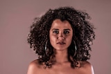 Kirsty (22) woman with natural curly hair and hoop earrings stares directly at viewer. For a story about black women's hair