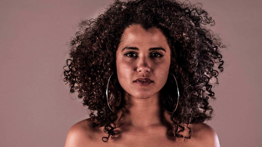 Kirsty (22) woman with natural curly hair and hoop earrings stares directly at viewer. For a story about black women's hair