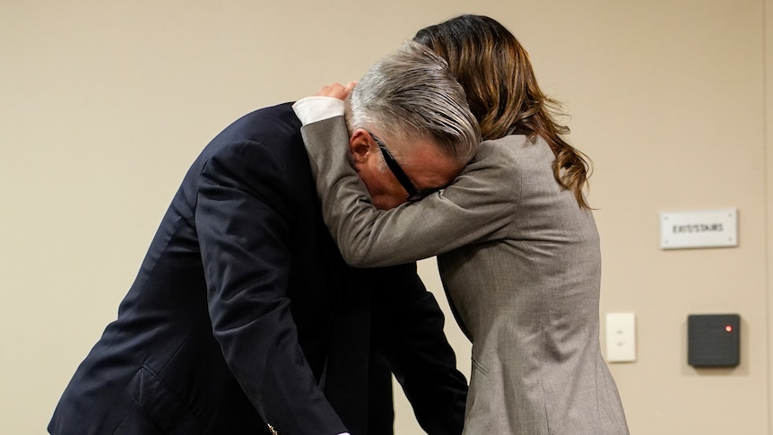 A man in a suit leans down while being embraced by a brunette woman in a grey suit, sombre 