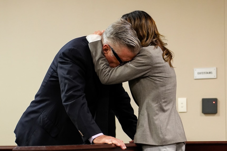 A man in a suit leans down while being embraced by a brunette woman in a grey suit, sombre 