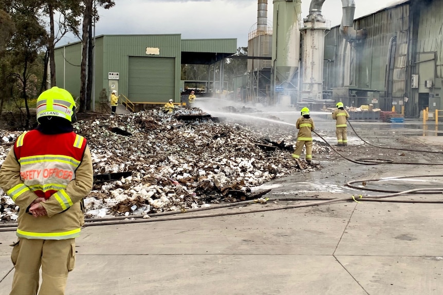 Two firefighters spray water of large piles of plastic bottles.