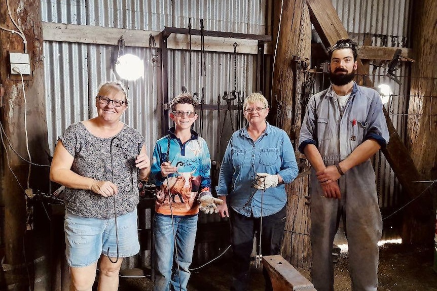 Four people standing side by side in a blacksmiths workshop holding small metal hooks.