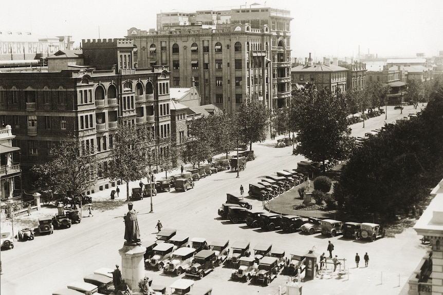 A black and white photo of buildings, parked cars and people looking west down North Terrace