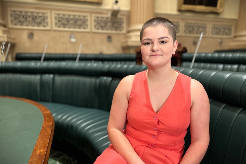 A young woman with shaved brown hair wearing a bright pink vest sitting on a bench inside South Australia's Parliament House.