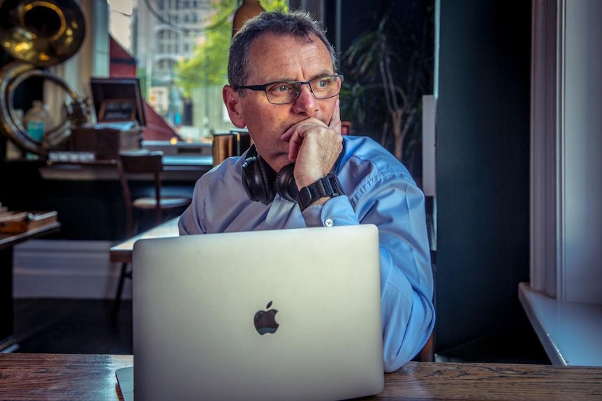 Man in a blue business shirt sitting at a desk with a laptop in front of him, looking thoughtful.