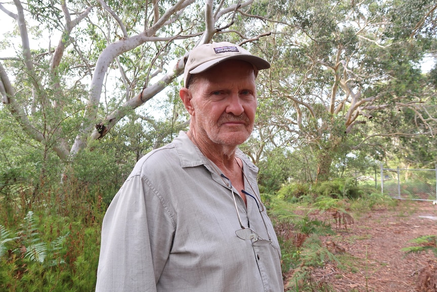 A man standing inside bushland with a hat and a grey button-up shirt stares at the camera.