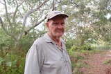 A man standing inside bushland with a hat and a grey button-up shirt stares at the camera.