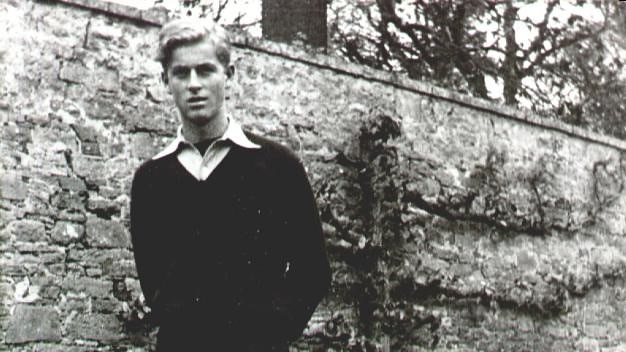 A black and white photograph of a young Prince Philip in dark shorts, dark jumper and white shirt.