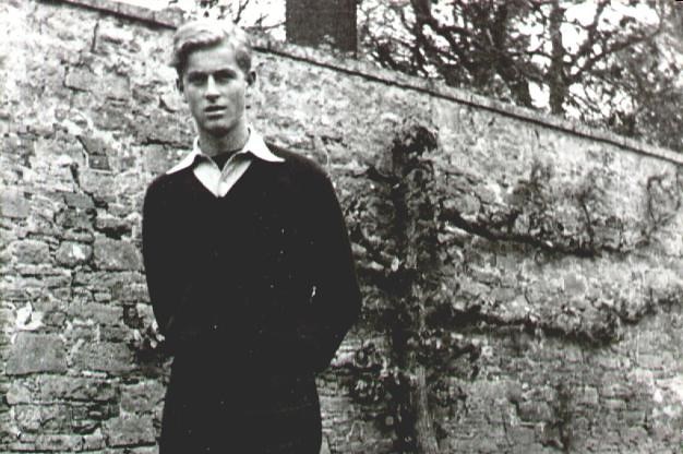 A black and white photograph of a young Prince Philip in dark shorts, dark jumper and white shirt.