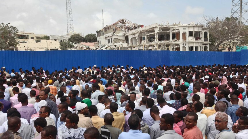 A huge crowd of people stand behind a blue fence. There are several destroyed buildings on the other side of the fence.