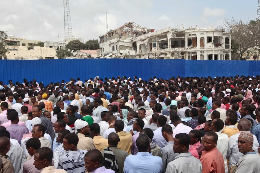 A huge crowd of people stand behind a blue fence. There are several destroyed buildings on the other side of the fence.