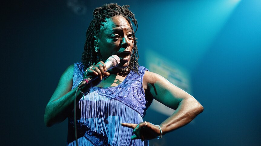 a woman with dreadlocks wearing a blue dress  sings into a microphone on stage