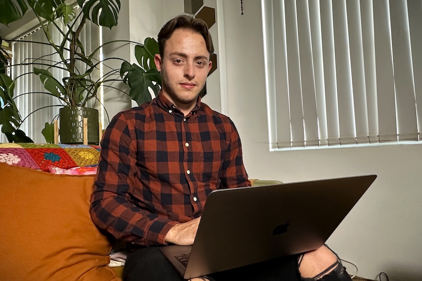 A man in a red and black checked shirt sits on a red sofa with a laptop on his lap.