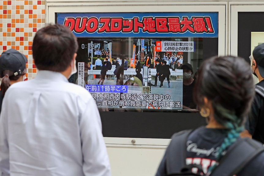 People stand facing a TV screen showing television reports that Shinzo Abe has been shot. 