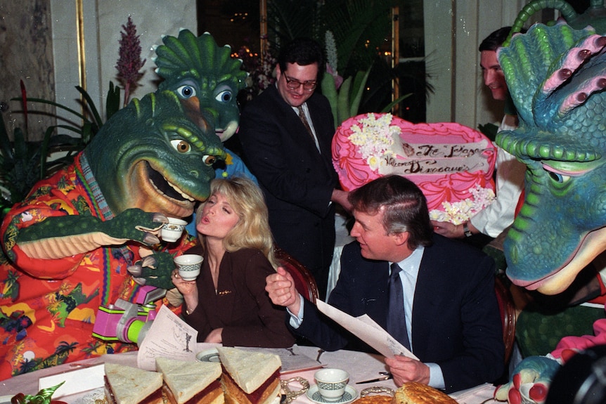 Trump sits at a table with Maples. The pair are surrounded by people dressed in lifesize dinosaur costumes. 