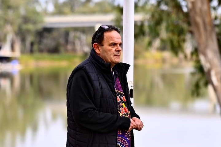 A man speaks in front of a big river. He wears a black jacket, a shirt with aboriginal artwork and black sunglasses