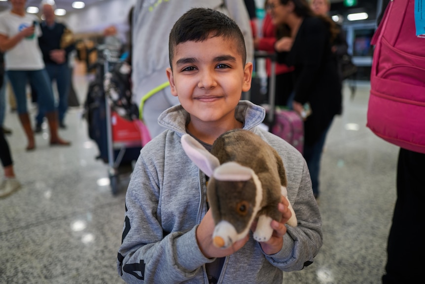 a young boy smiling holding a stuffed toy at the arrivals section of the airport