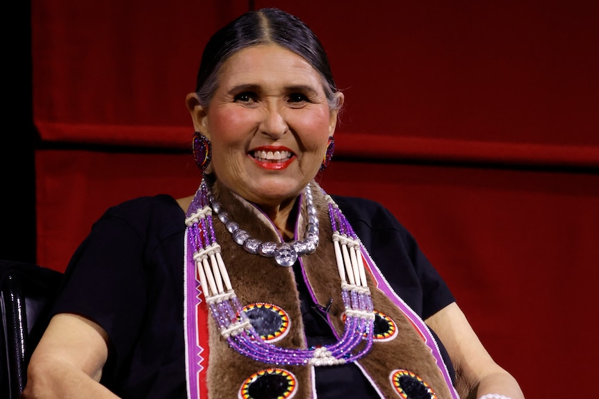 A smiling woman sits on stage wearing some traditional Native American garments.