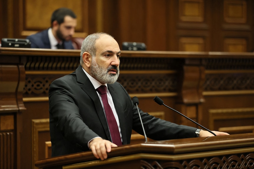 Man in black suit speaks at wooden podium in parliament.